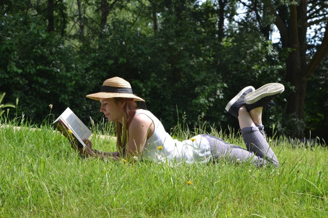 woman lying in grass on stomach, wearing straw hat and reading a book